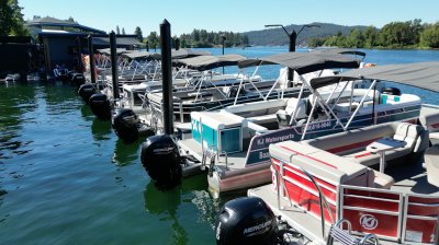 Boats docked on a sunny lake with trees and hills in the background.